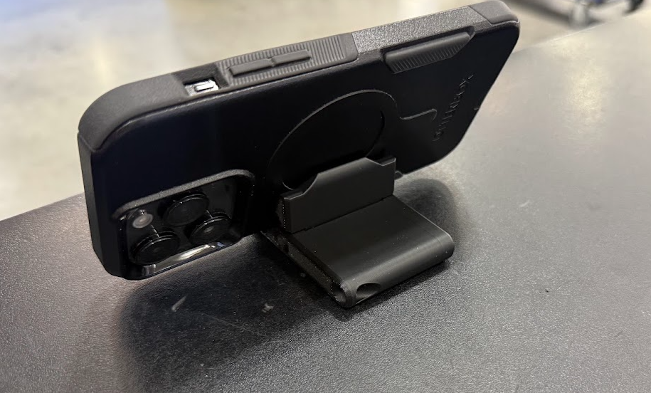 Black 3D printed foldable phone stand holding a phone at an angle on a desk